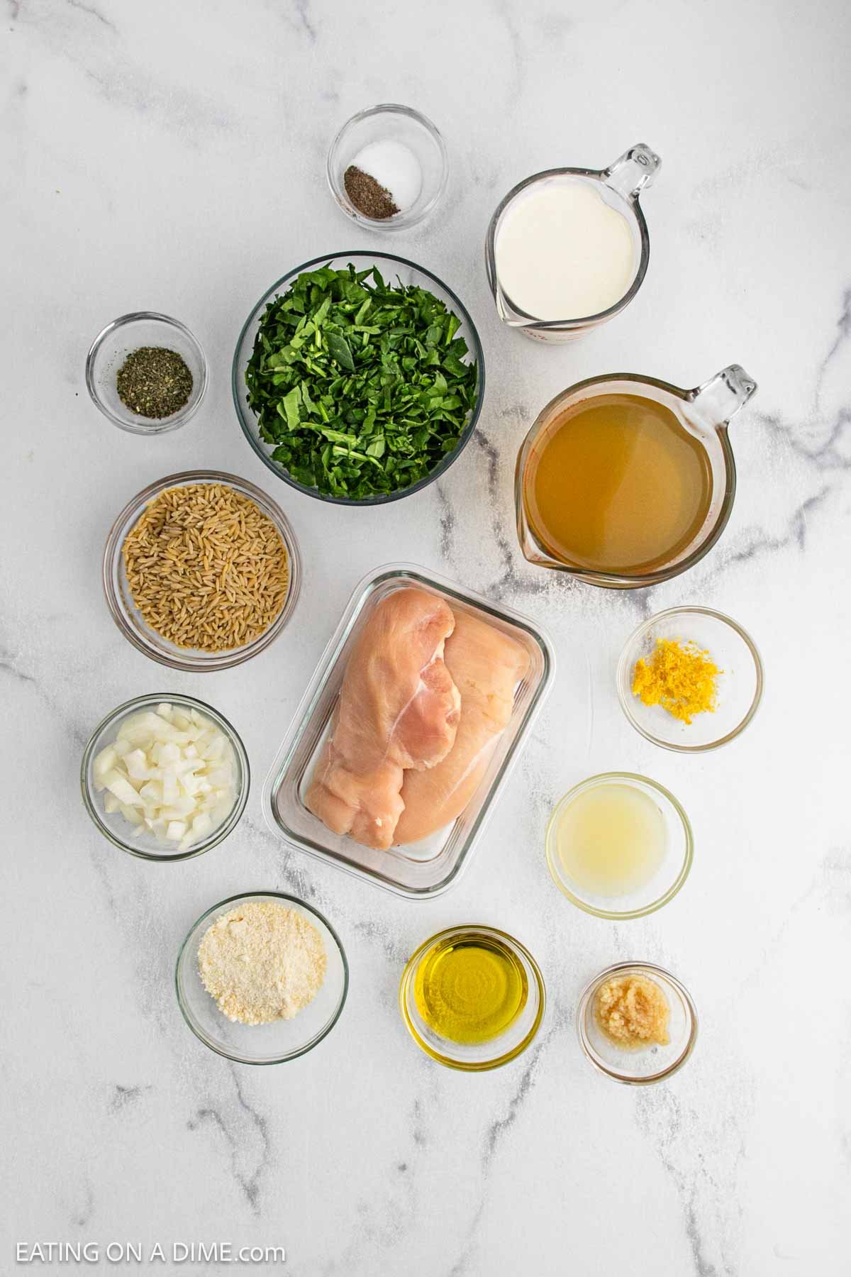 Overhead view of ingredients for Lemon Chicken Orzo in bowls on a white surface, including raw chicken breasts, orzo pasta, chicken broth, lemon zest and juice, olive oil, parmesan, spinach, onion, garlic, dried herbs, milk, and pepper.