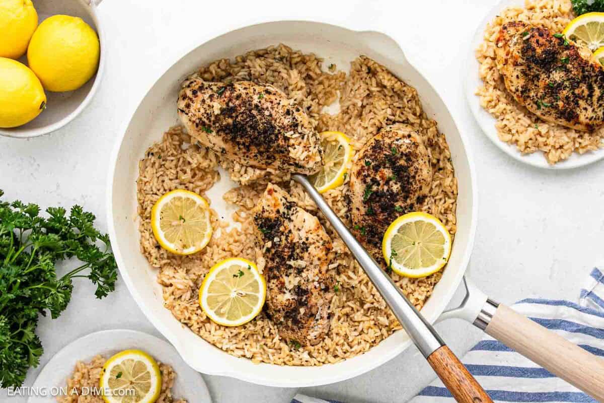 A white skillet filled with seasoned Lemon Chicken and Rice, garnished with lemon slices. A serving is plated nearby, with fresh parsley and whole lemons also visible on the table.