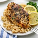 A plate with a seasoned, baked Lemon Chicken breast on a bed of rice, garnished with fresh parsley and two lemon slices. A fork rests on the side of the plate, beside a blue-striped napkin—perfect for a classic Chicken and Rice meal.
