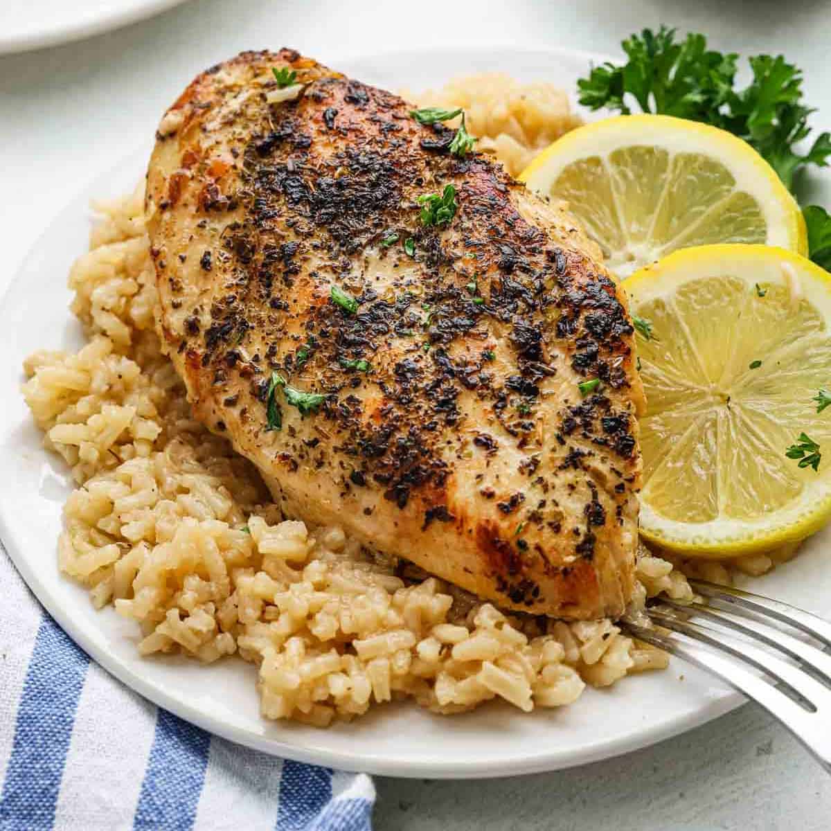 A plate with a seasoned, baked Lemon Chicken breast on a bed of rice, garnished with fresh parsley and two lemon slices. A fork rests on the side of the plate, beside a blue-striped napkin—perfect for a classic Chicken and Rice meal.