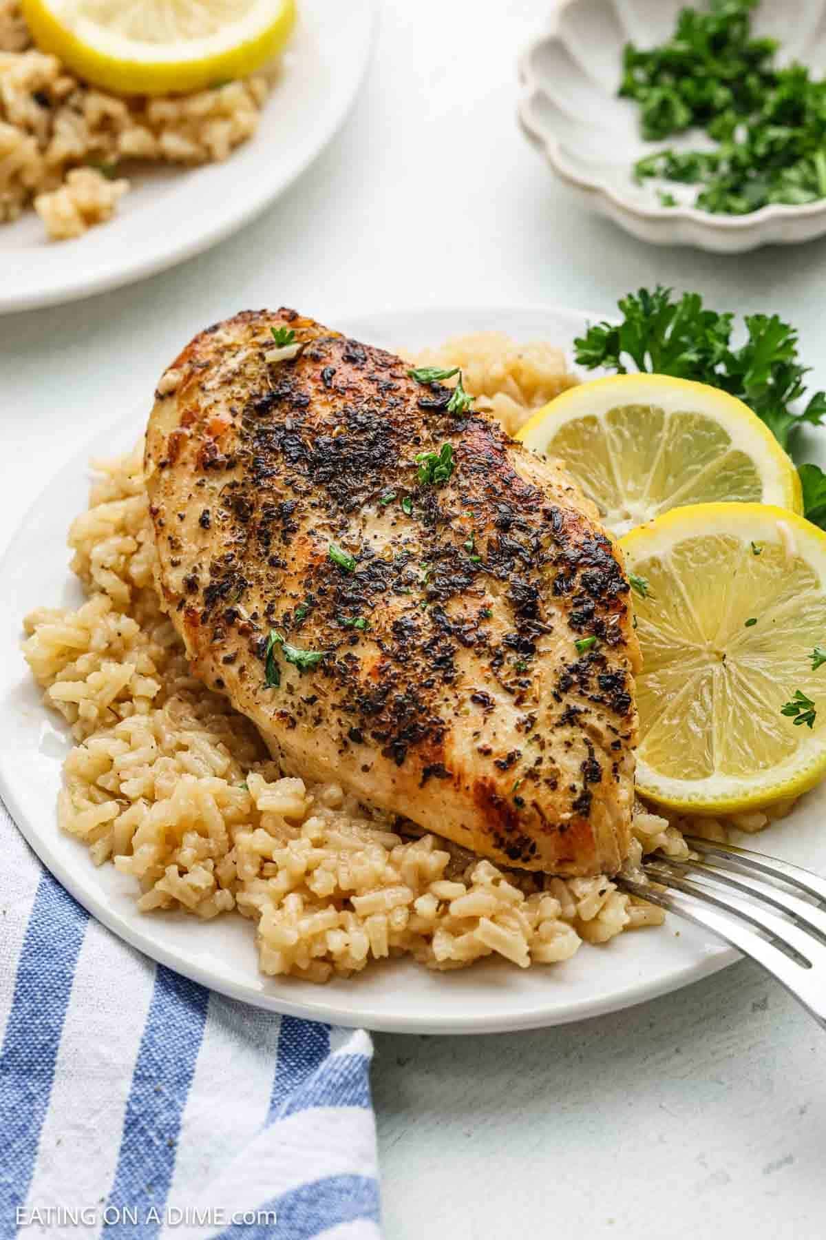 A seasoned baked Lemon Chicken breast sits on a bed of rice, garnished with fresh parsley and lemon slices, served on a white plate with a fork and a blue and white striped napkin nearby—a classic Lemon Chicken Recipe.