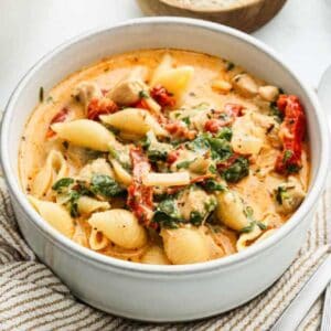 A white bowl filled with creamy soup containing pasta shells, pieces of chicken, spinach, sun-dried tomatoes, and herbs sits atop a beige-striped cloth—perfect for adding comfort to your Meal Plan 96.