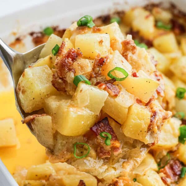A close-up of a spoonful of cheesy, baked Mississippi Mud Potatoes with bits of bacon and sliced green onions, held above a casserole dish filled with more of the hearty potato mixture.