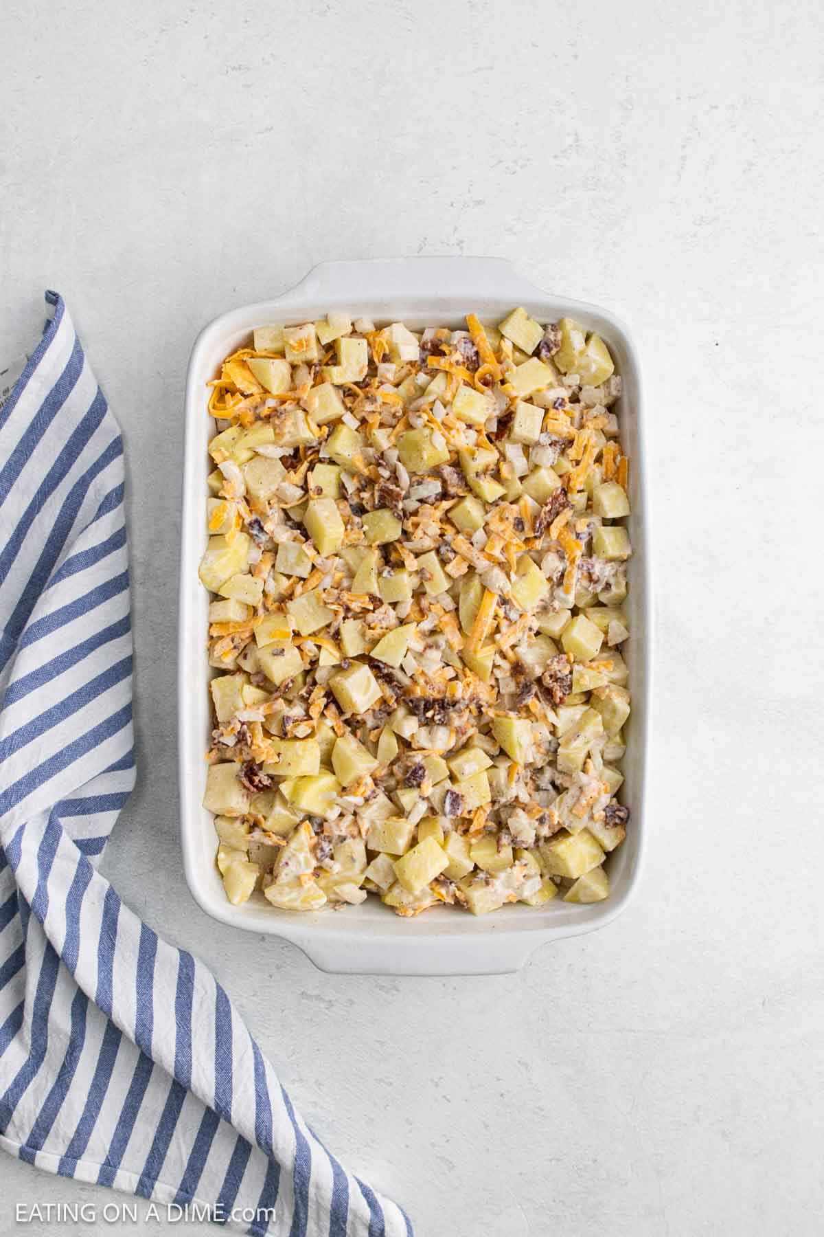 A casserole dish filled with cheesy potatoes, bacon bits, and melty cheese, set on a light surface next to a blue and white striped kitchen towel—classic Mississippi Mud Potatoes at their best.