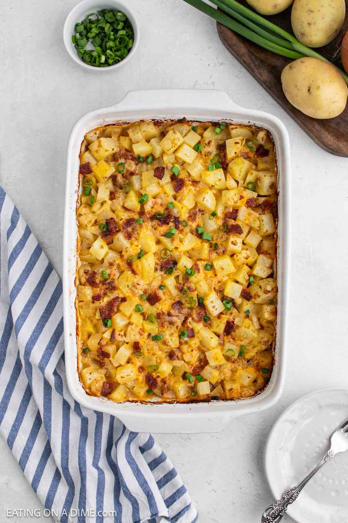 A baked potato casserole dish filled with cheesy potatoes, topped with crispy bacon bits and chopped green onions, sits next to a bowl of sliced green onions, whole potatoes, and a striped kitchen towel—classic Mississippi Mud Potatoes.