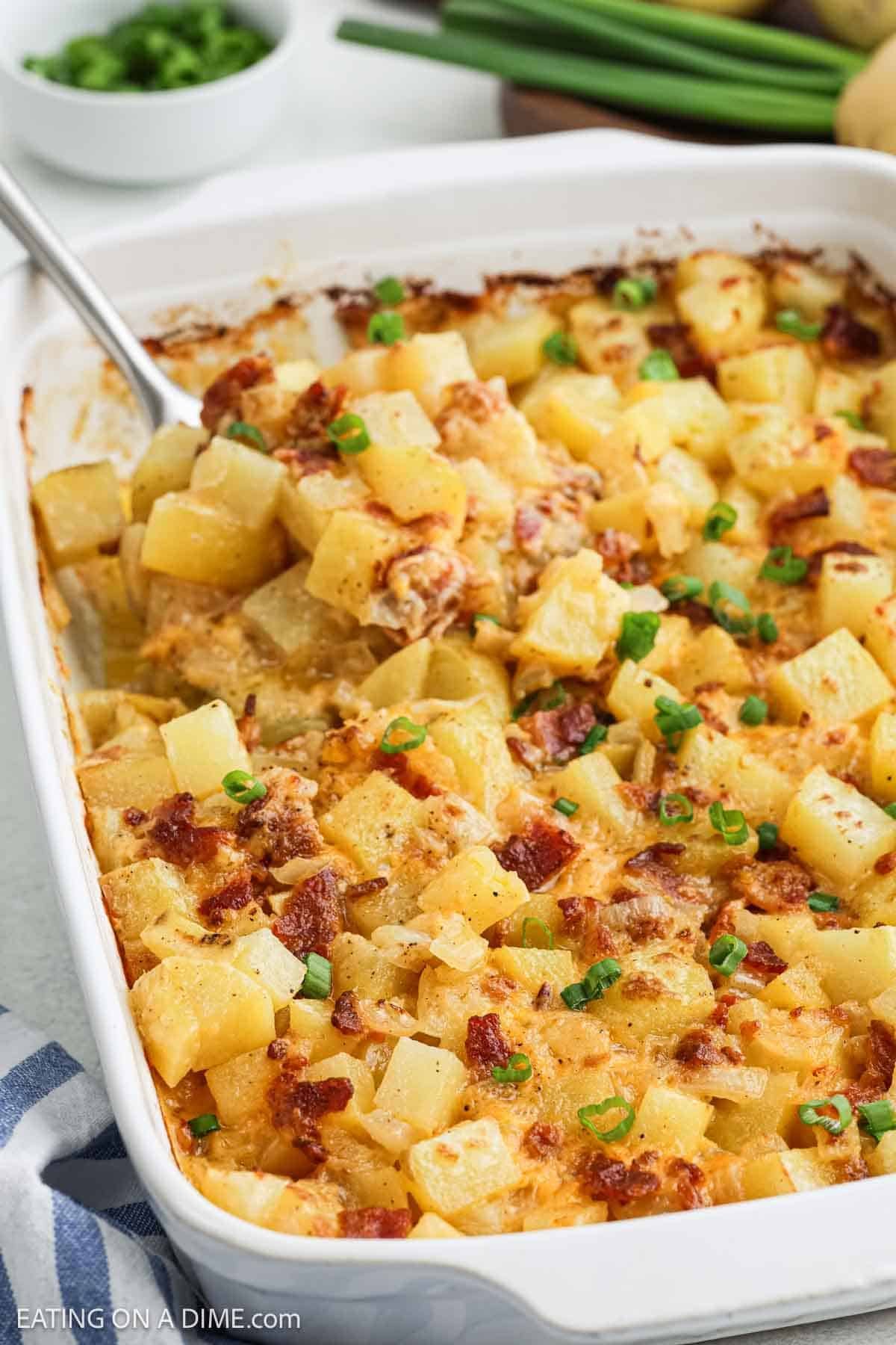 A casserole dish of cheesy potatoes, known as Mississippi Mud Potatoes, is topped with crispy bacon bits and chopped green onions. A serving spoon scoops out this rich potato casserole, with fresh green onions in a bowl nearby.