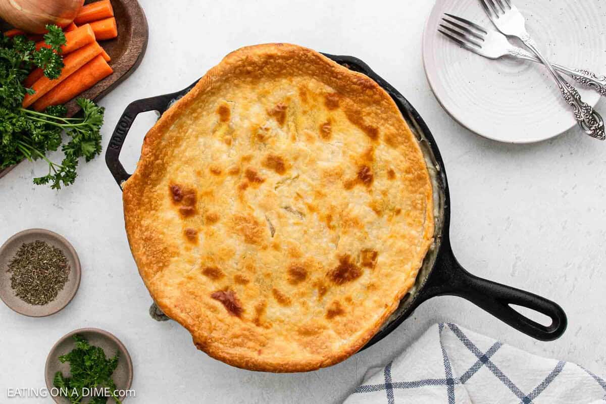 A golden-brown skillet chicken pot pie in a cast iron skillet sits on a white surface, surrounded by fresh parsley, carrot sticks, a bowl of herbs, an empty plate with forks, and a towel.