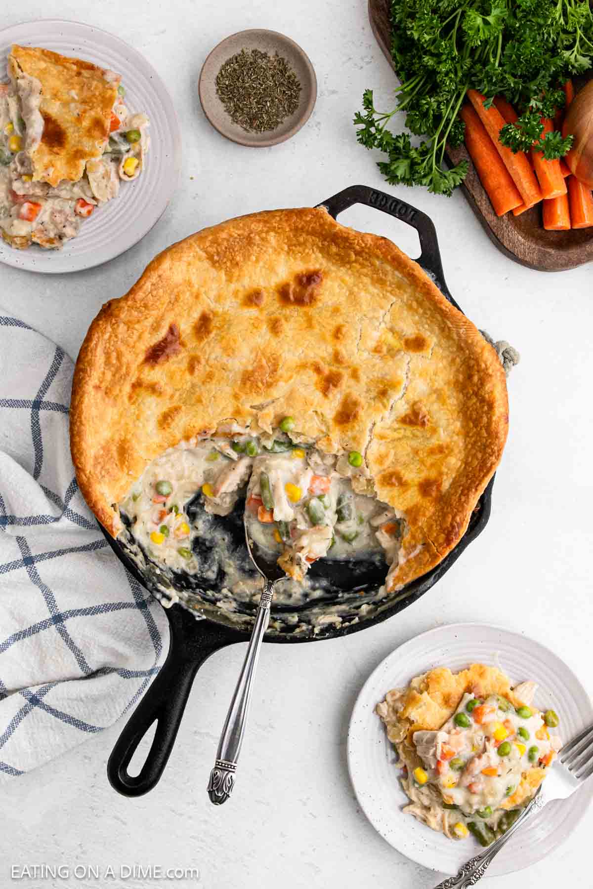 A cast iron skillet filled with Skillet Chicken Pot Pie, its creamy filling topped with golden-brown crust. Nearby, two plates hold portions of the pie, surrounded by fresh parsley, carrots, and a bowl of pepper.