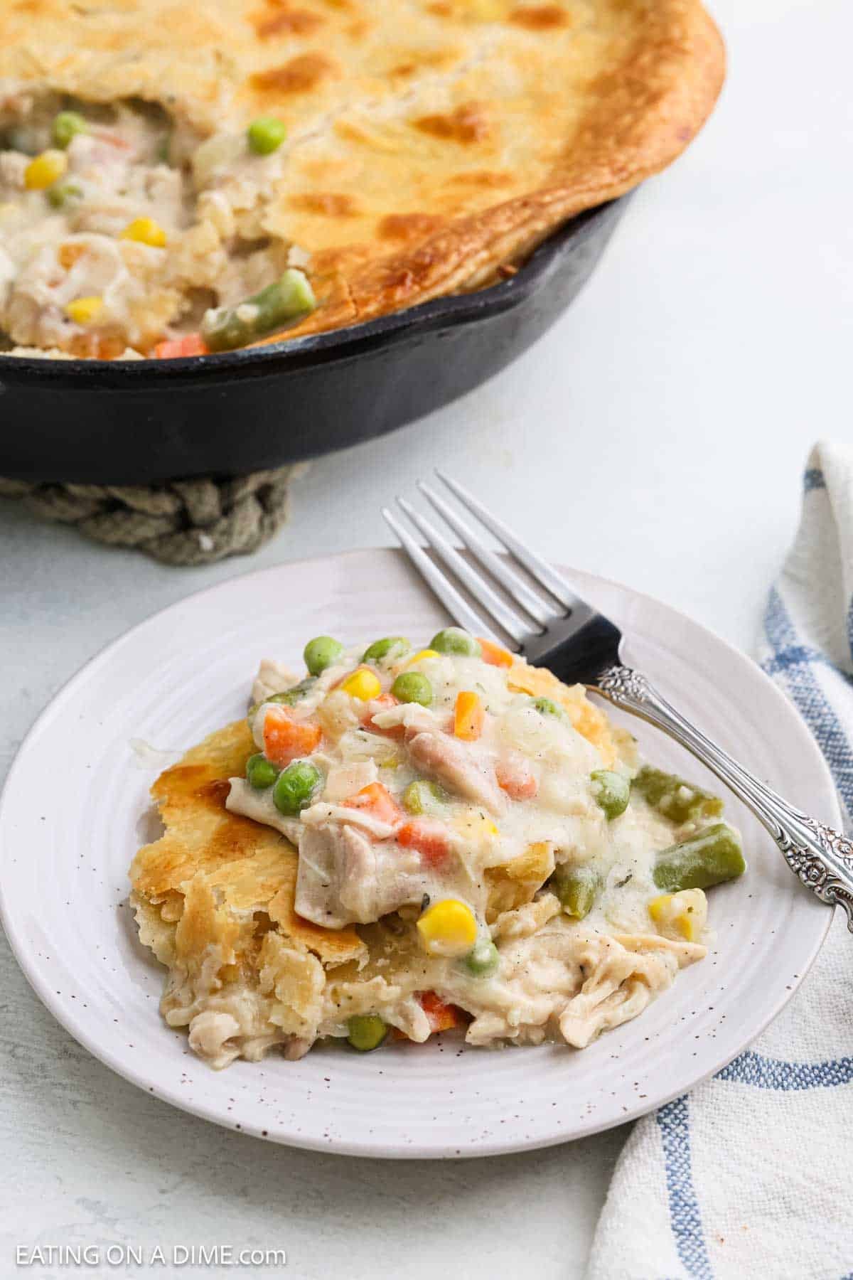 A serving of creamy Skillet Chicken Pot Pie with mixed vegetables and a golden crust on a white plate, with a fork beside it. The remaining pot pie sits in a cast iron skillet in the background.