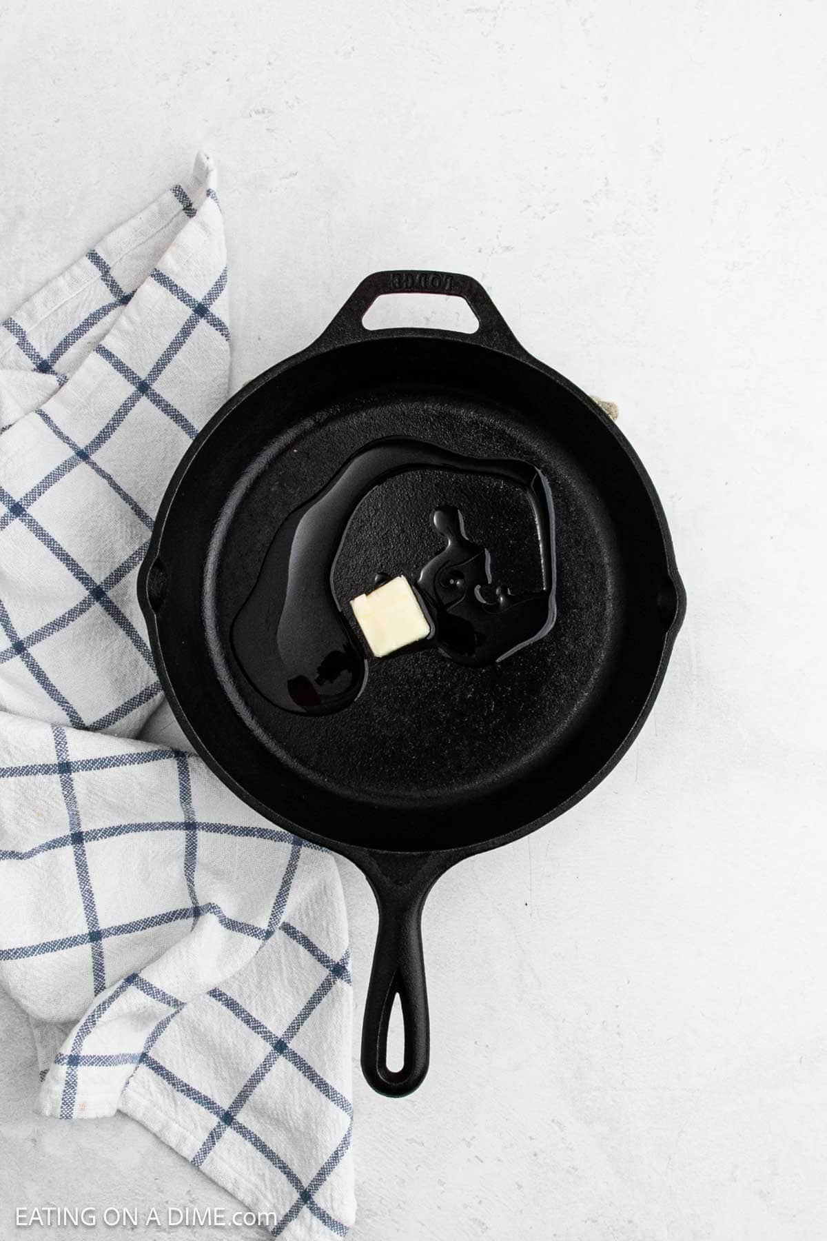 A black cast iron skillet with a pat of butter and melted butter inside sits on a white surface next to a blue and white checkered kitchen towel, ready to start your favorite Skillet Chicken Pot Pie recipe.