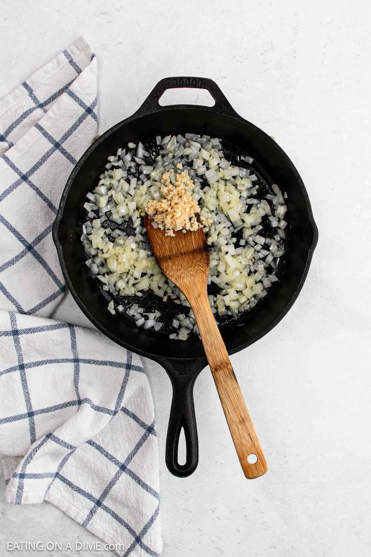 A cast iron skillet with chopped onions and minced garlic being sautéed, stirred with a wooden spatula—perfect beginnings for skillet recipes like Skillet Chicken Pot Pie. A blue and white checkered towel lies beside the skillet on a white surface.