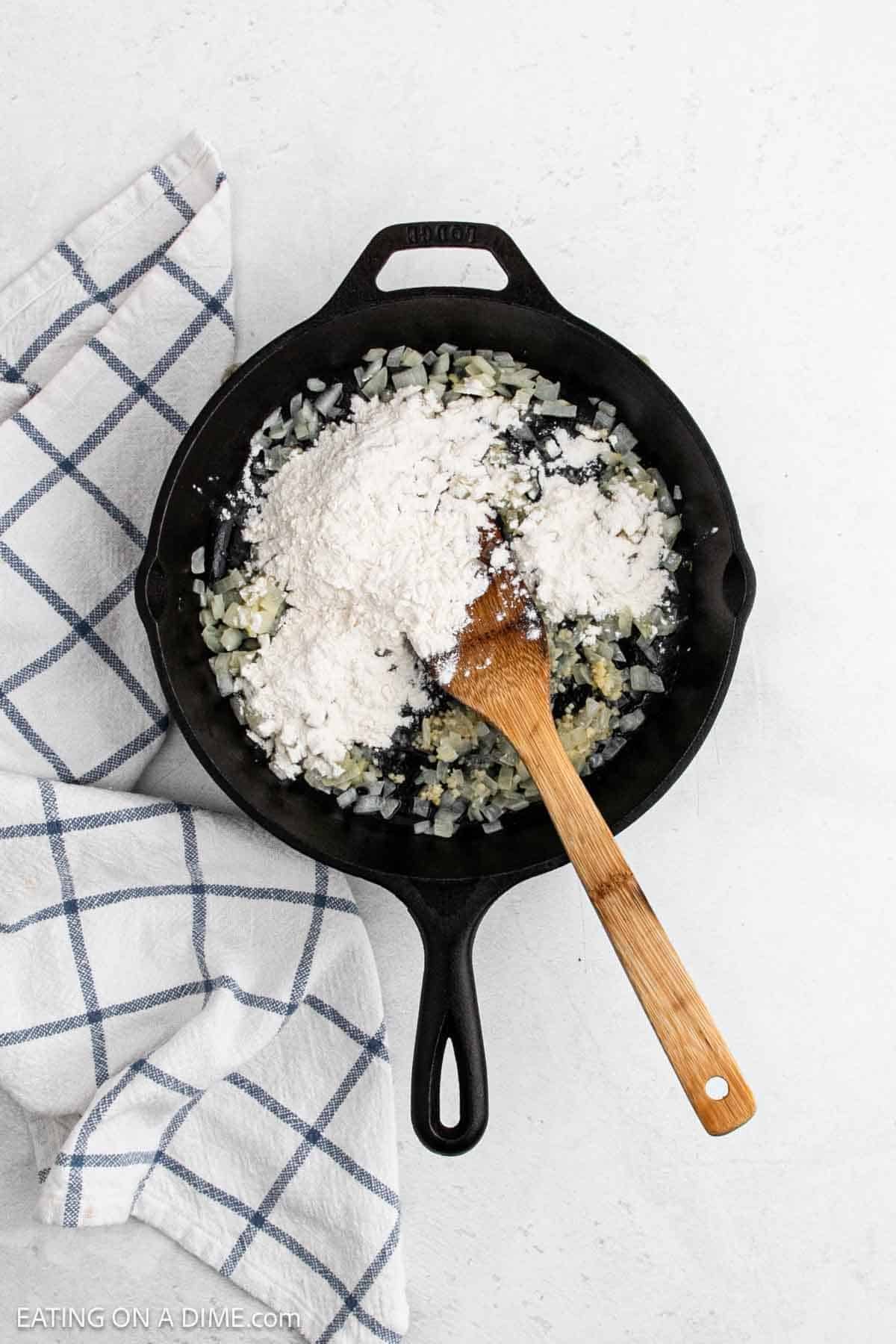 A black cast iron skillet with diced onions and flour being stirred by a wooden spoon, perfect for starting a Skillet Chicken Pot Pie, sits on a white surface next to a white and blue checkered kitchen towel.