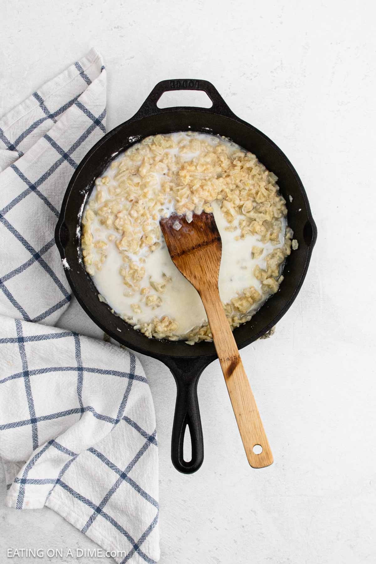 A cast iron skillet filled with creamy oats and milk sits on a white surface, with a wooden spatula resting inside. Perfect for trying a new skillet recipe, a white and blue checkered kitchen towel is beside the skillet.