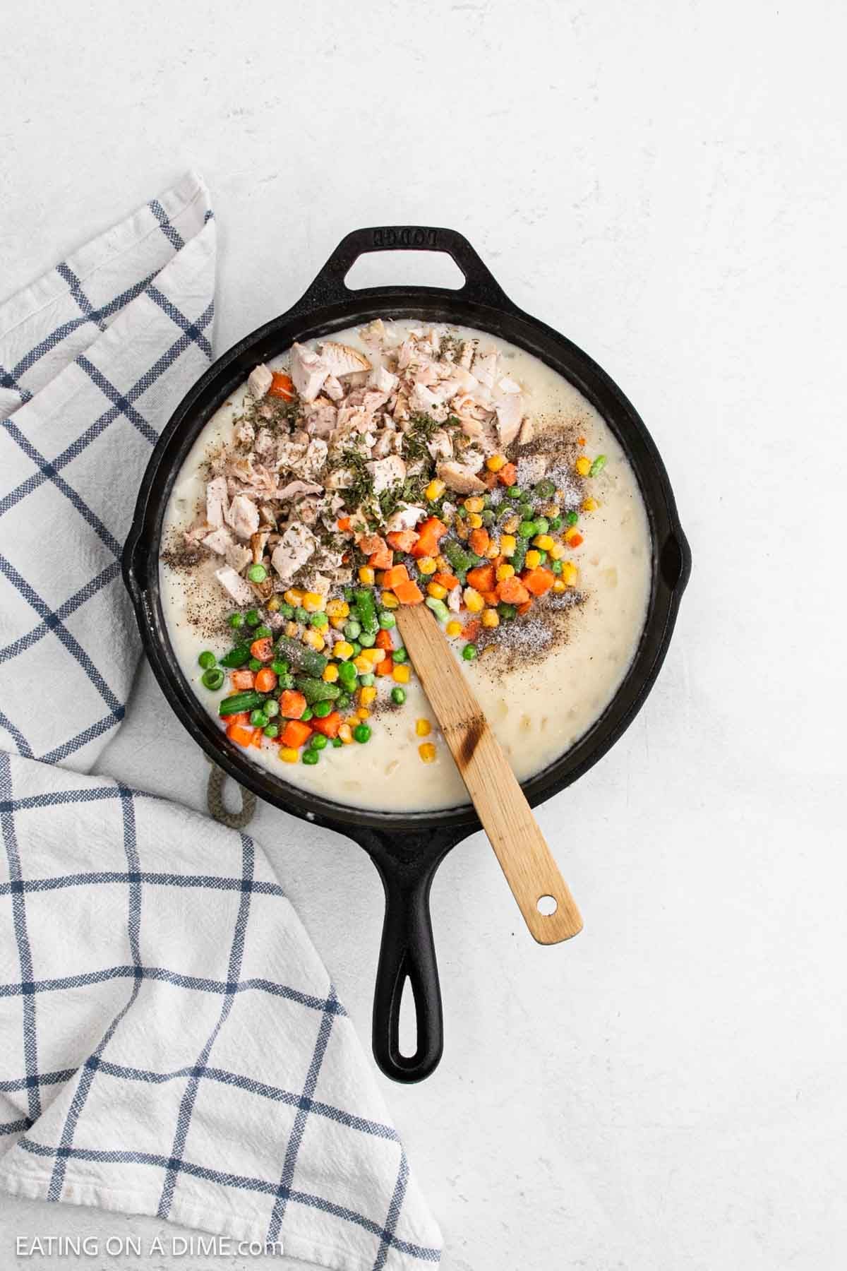 A black skillet filled with a creamy chicken pot pie mixture, topped with chopped cooked chicken, peas, carrots, and green beans. A wooden spoon rests in the skillet on a white surface next to a blue-striped kitchen towel.