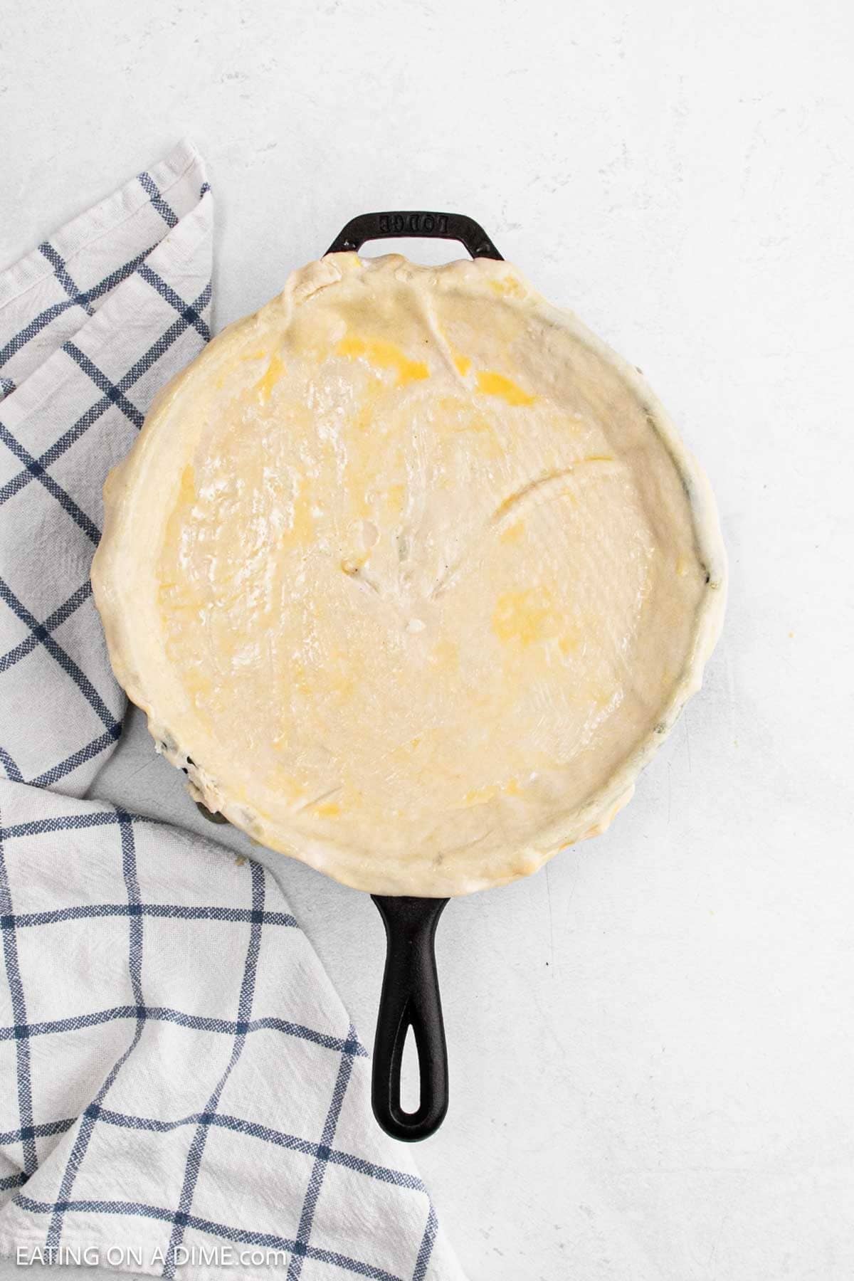 A round, unbaked Skillet Chicken Pot Pie with a golden egg wash rests in a black cast iron skillet. A blue and white checkered kitchen towel is partially visible beside the skillet on a white surface.