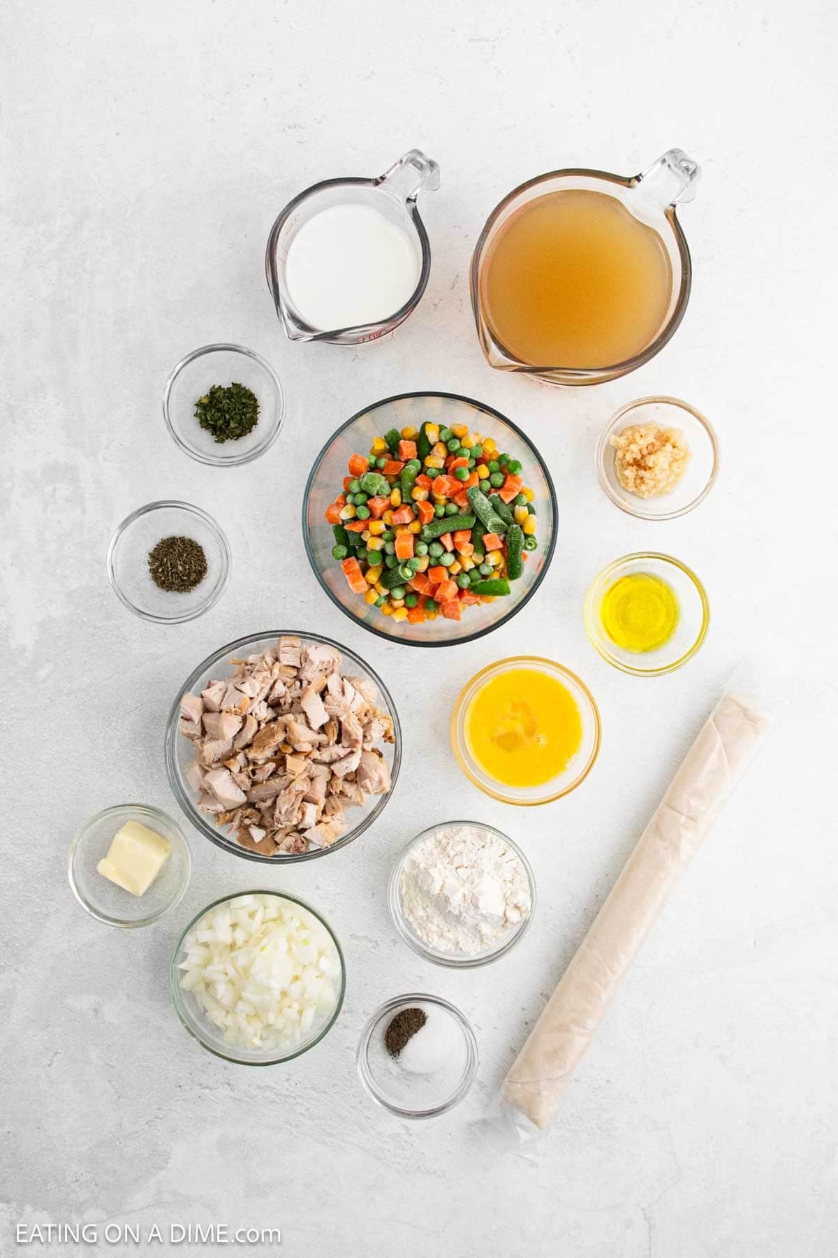 Overhead view of Skillet Chicken Pot Pie ingredients on a white surface, including milk, broth, mixed vegetables, garlic, olive oil, seasonings, chopped cooked chicken, melted butter, flour, pie crust, onions, and black pepper.