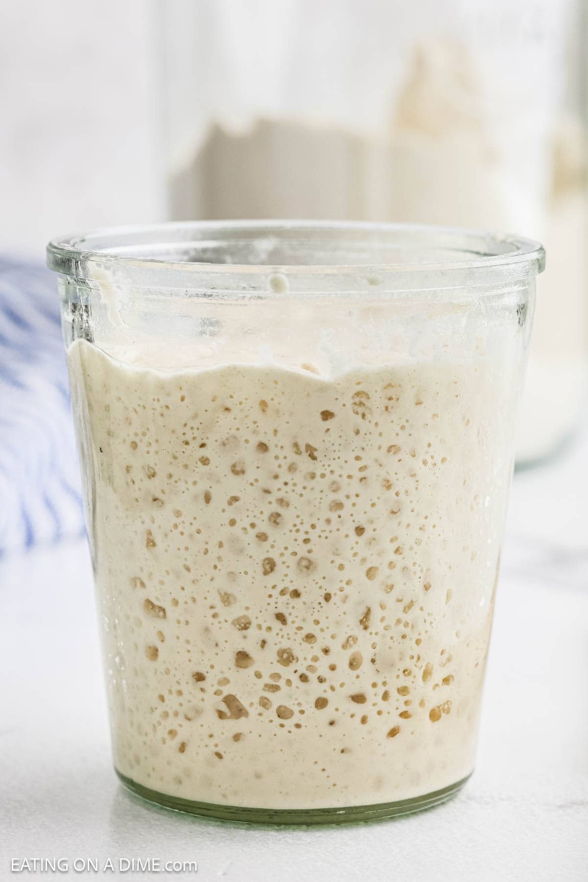 A glass jar filled with bubbly, active sourdough starter sits on a white surface, perfect for anyone learning how to make sourdough, with another jar blurred in the background.