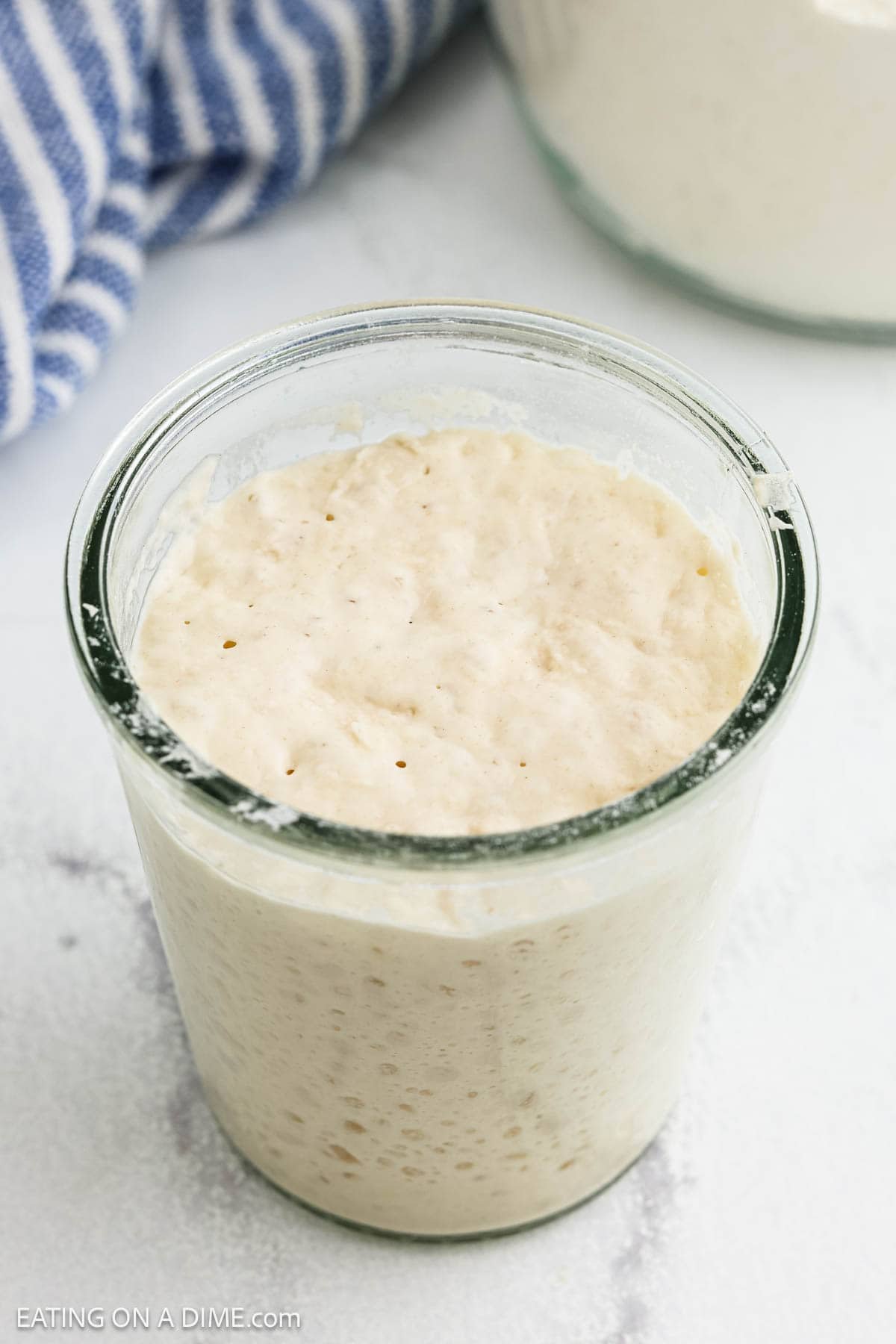 A glass jar filled with bubbly, active sourdough starter sits on a light surface, perfect for anyone wanting to learn how to make sourdough, with a blue-striped towel and another jar in the background.