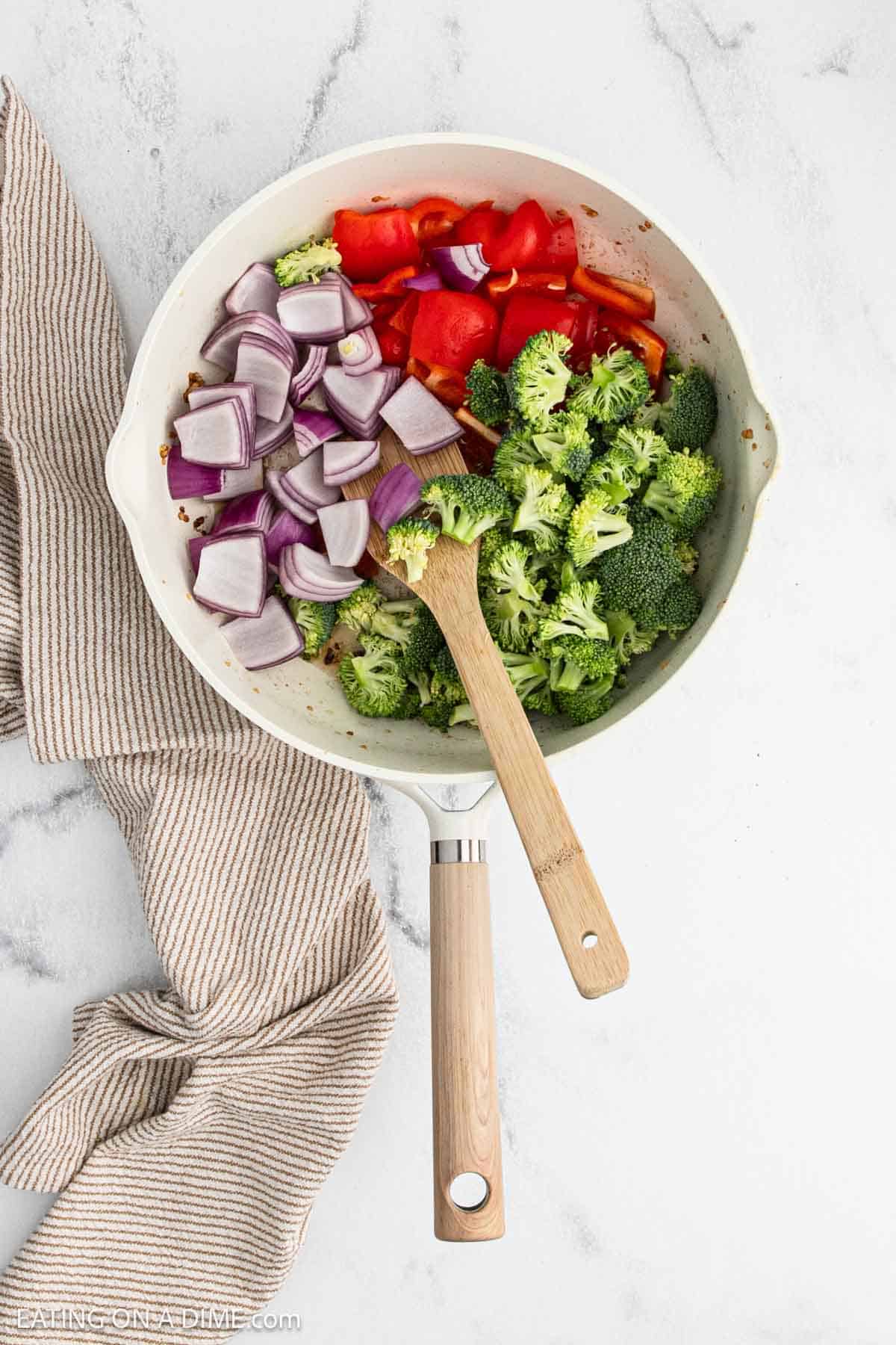Fresh red onion, red bell pepper, and chopped broccoli in a large skillet with a wooden spatula.