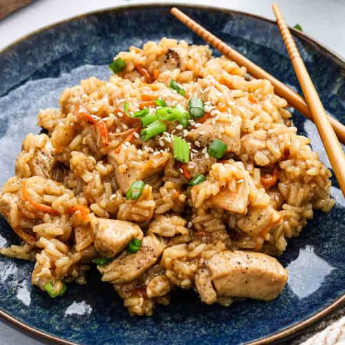 A plate of teriyaki chicken & rice fried together, garnished with chopped green onions and sesame seeds, served with a pair of wooden chopsticks on the side.