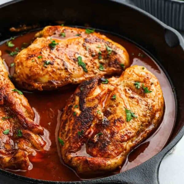 Three seasoned, golden-brown chicken breasts sizzle in a cast iron skillet with a rich, reddish-brown sauce and fresh herbsโan irresistible highlight from Meal Plan 97.