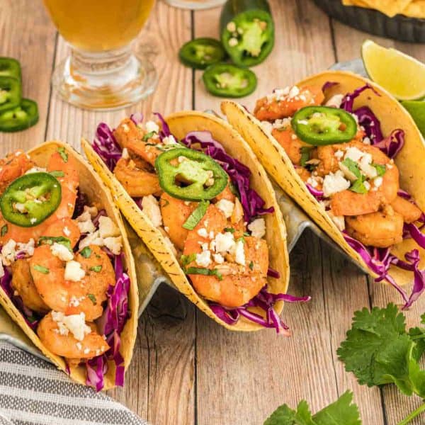 Three shrimp tacos topped with sliced jalapeรฑos, crumbled cheese, and cilantro, served on corn tortillas with shredded red cabbage. Perfect for Meal Plan 95. Lime wedges, cilantro, and a drink are in the background on a wooden table.
