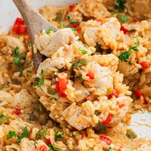 A close-up of a wooden spoon lifting a serving of Cajun Chicken and Rice mixed with chopped red and green bell peppers, herbs, and seasonings from a pan.