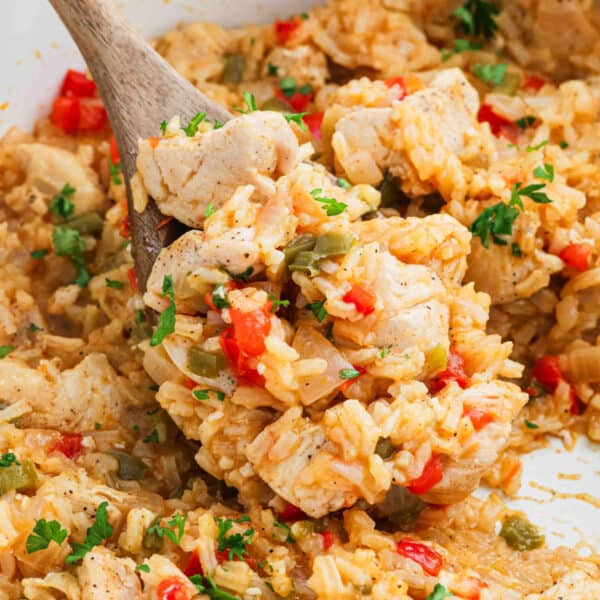 A close-up of a wooden spoon lifting a serving of Cajun Chicken and Rice mixed with chopped red and green bell peppers, herbs, and seasonings from a pan.