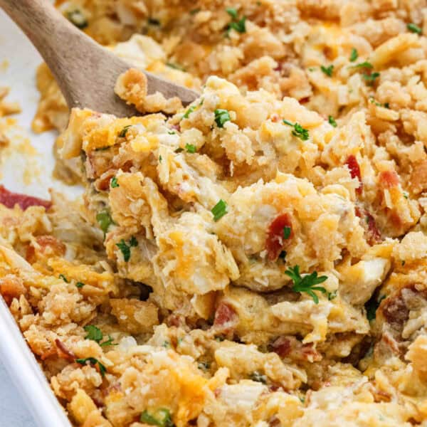 A close-up of a creamy, cheesy Neiman Marcus Chicken Casserole with a crunchy topping being scooped out with a wooden spoon. The dish contains visible bits of vegetables and is garnished with fresh chopped herbs.