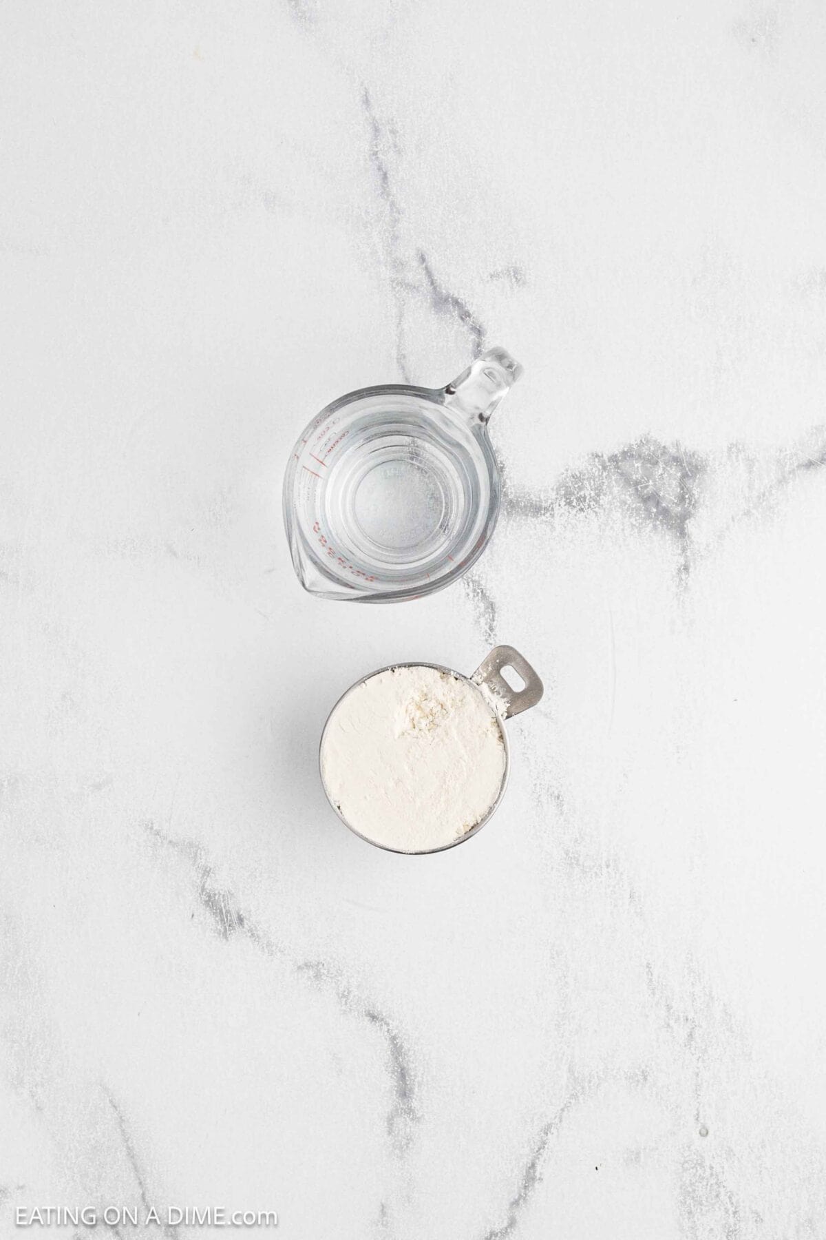 A glass measuring cup filled with water and a metal measuring cup filled with flour are placed on a white marble surface, ready to make sourdough or prepare your favorite sourdough recipe.