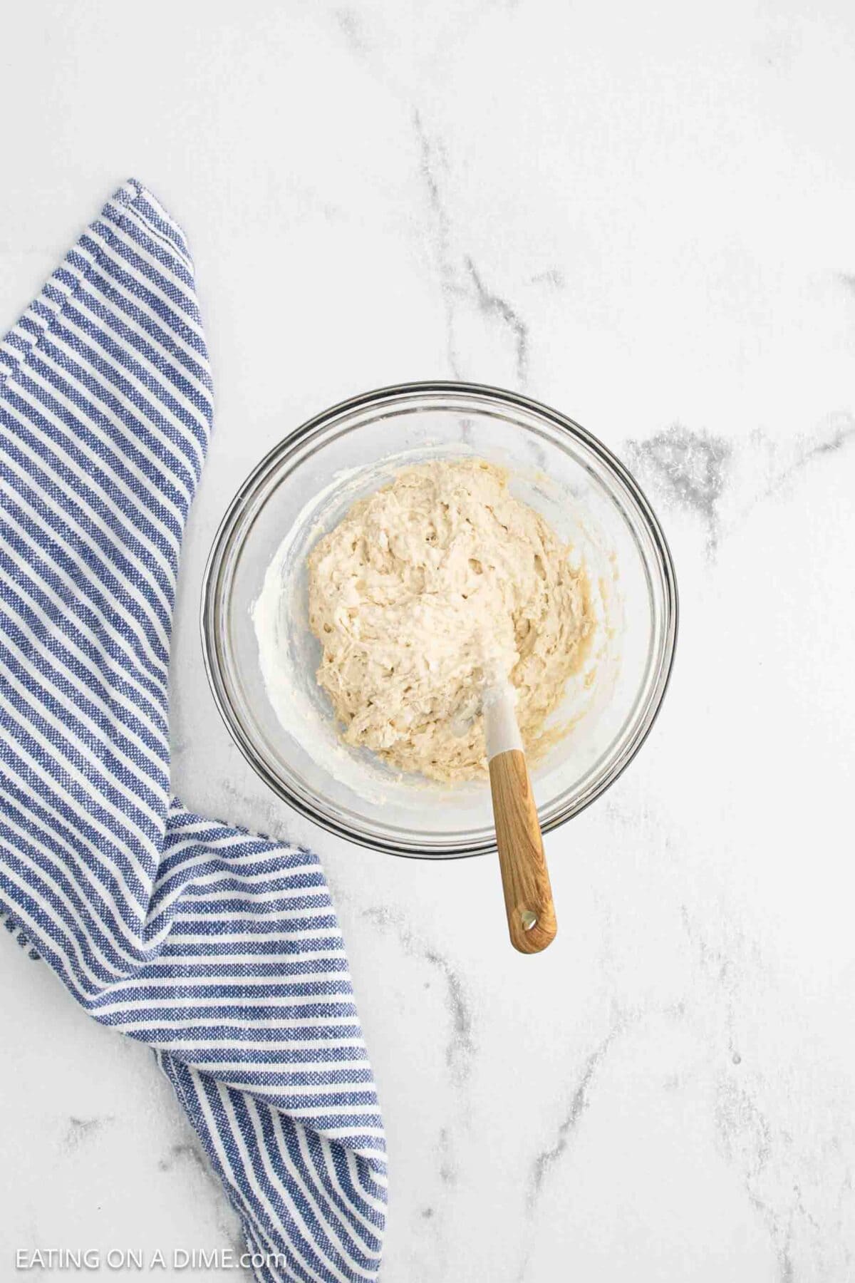 A glass bowl with sourdough starter bread dough and a wooden-handled spatula sits on a white marble surface next to a blue and white striped kitchen towel, ready for your next sourdough recipe.