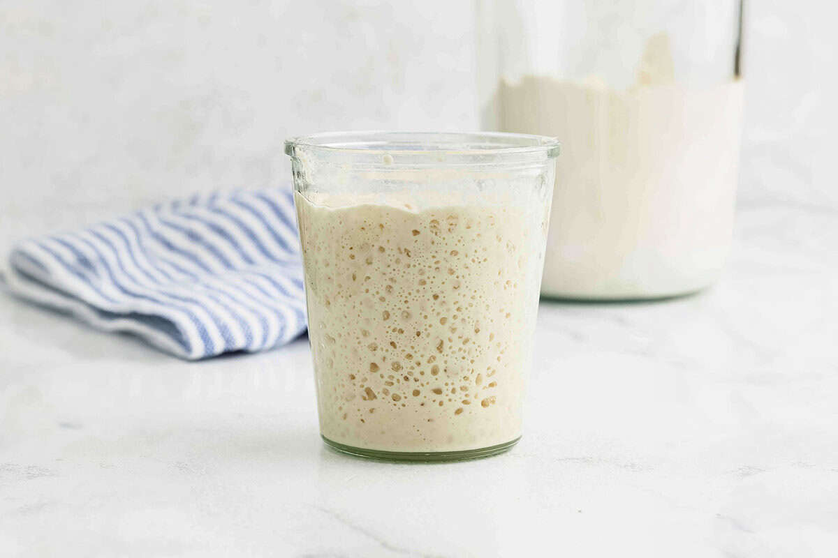 A glass jar filled with bubbly, active sourdough starter sits on a white countertop—a perfect example of How to Make a Sourdough Starter. In the background, a blue and white striped cloth and a larger jar partially filled with flour add charm.
