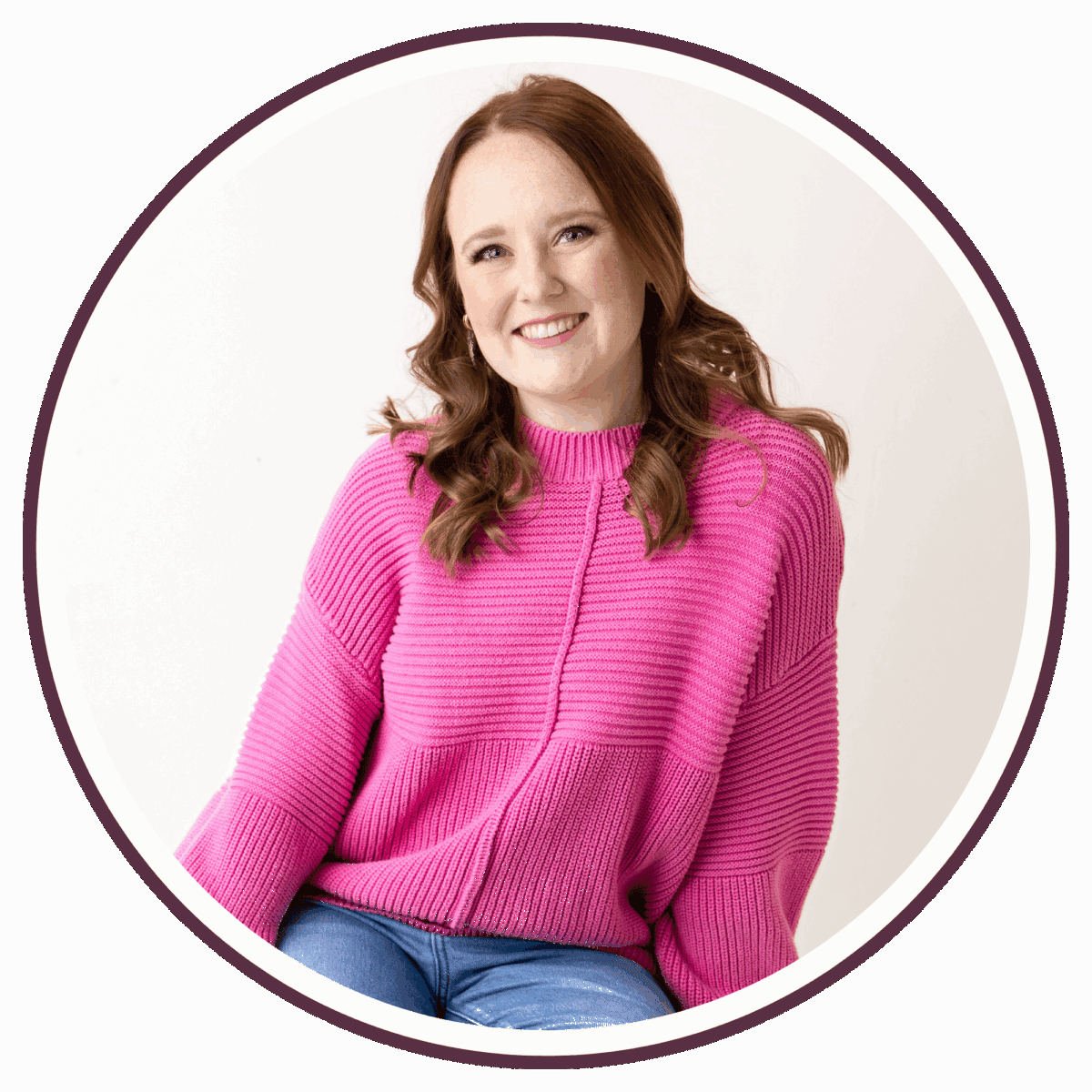 Carrie, a woman with fair skin and long auburn hair, wearing a bright pink sweater and blue jeans, smiles while sitting against a white background inside a circular frame.