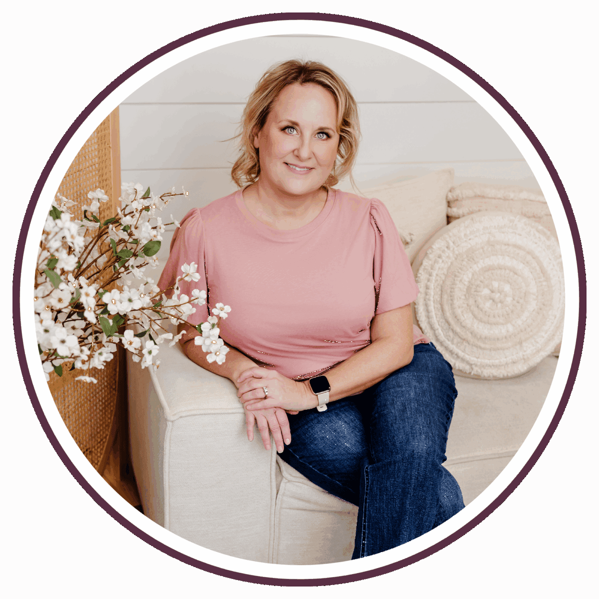 Carrie, a woman with blonde hair, wearing a pink shirt and blue jeans, sits on a cream-colored couch next to a pillow and white flowers, smiling gently at the camera.