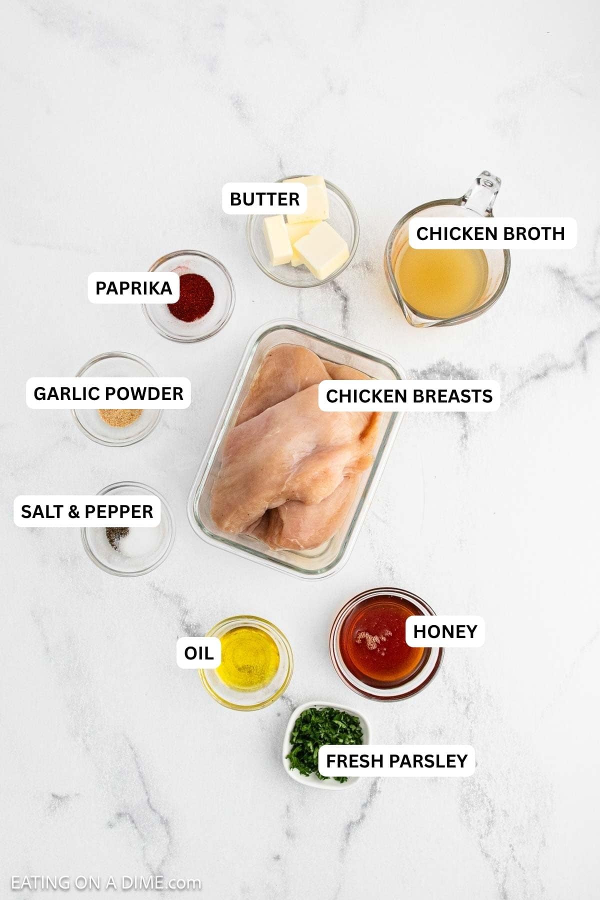 Ingredients for recipe: Raw chicken breasts in a glass container surrounded by small bowls of butter, chicken broth, paprika, garlic powder, pepper, olive oil, hot sauce, and chopped parsley.
