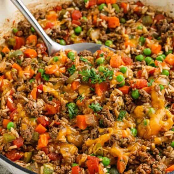 A close-up of a skillet filled with ground beef, rice, peas, diced tomatoes, carrots, green beans, onions, and melted cheddar cheeseโan ideal Meal Plan 99 optionโgarnished with fresh parsley and a serving spoon in the pan.