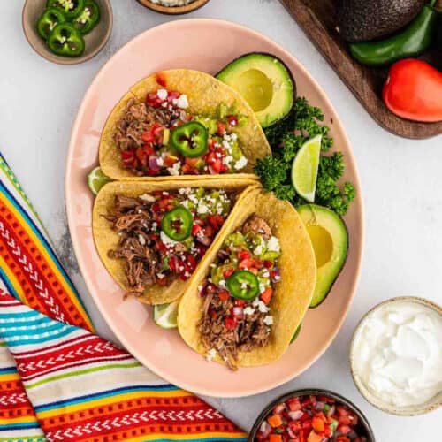 3 Pulled Pork Tacos topped with sliced jalapenos and pico de gallo with a side of avocado.