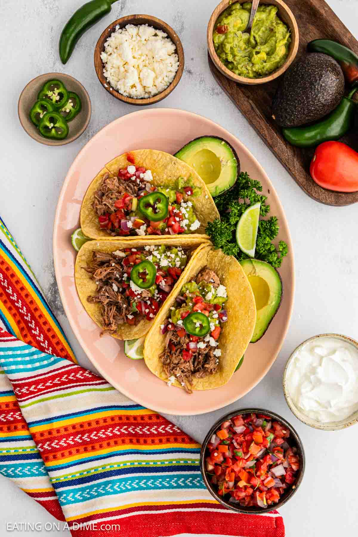 3 Shredded Pork Tacos on a large platter with a side of avocados with bowls of pico de gallo, sour cream, slice jalapenos, and guacamole on the side.