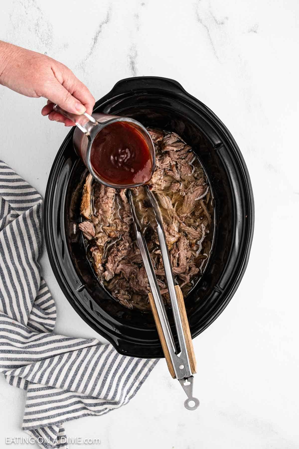 Shredded pork into the crock pot with BBQ sauce being poured over the pork.