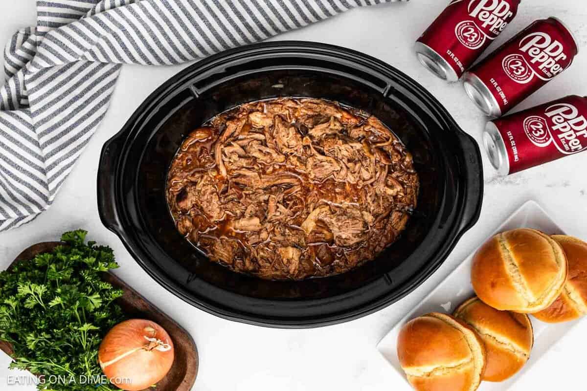 Shredded pulled pork mixed with juices in the slow cooker with a side of Dr Pepper Cans and hamburger buns.