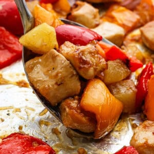 A close-up of a spoonful of Hawaiian Chicken Sheet Pan, featuring chunks of chicken, red and yellow bell peppers, and pineapple, all coated in a glossy sweet and sour sauce.