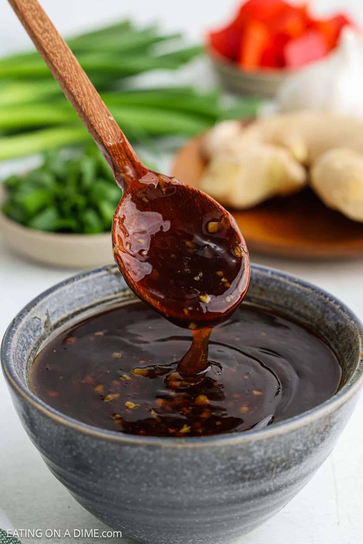 A wooden spoon lifts a thick, glossy teriyaki sauce from a ceramic bowl. Fresh green onions, ginger, and vegetables are blurred in the background.