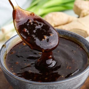 A spoon lifts thick, glossy homemade teriyaki sauce from a bowl, with fresh ginger and green onions blurred in the background.