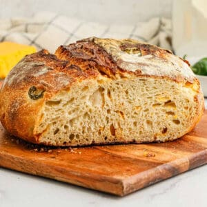A loaf of Jalapeño Cheddar Sourdough Bread with a golden, crispy crust and airy interior sits sliced in half on a wooden cutting board, its surface dotted with baked-in jalapeño pieces and melted cheddar.