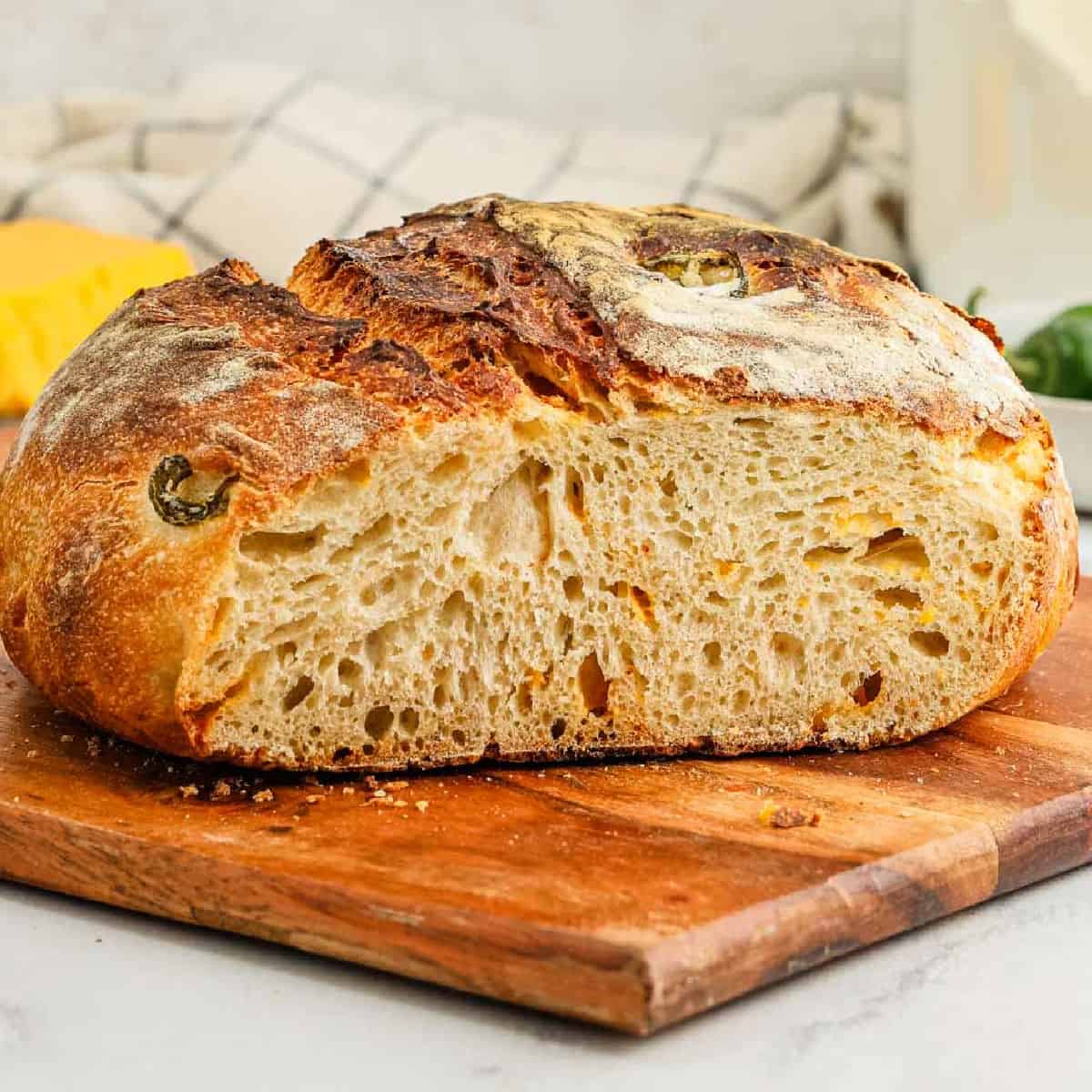 A loaf of Jalapeño Cheddar Sourdough Bread with a golden, crispy crust and airy interior sits sliced in half on a wooden cutting board, its surface dotted with baked-in jalapeño pieces and melted cheddar.