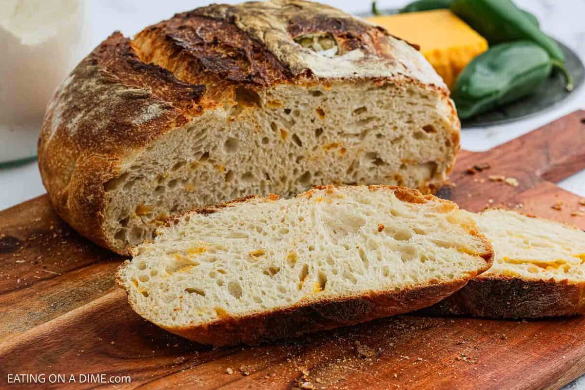A rustic Jalapeño Cheddar Sourdough Bread loaf with a dark, crispy crust sits on a wooden cutting board, two thick slices revealing a soft, airy interior. Cheese, jalapeños, and milk are visible in the background.