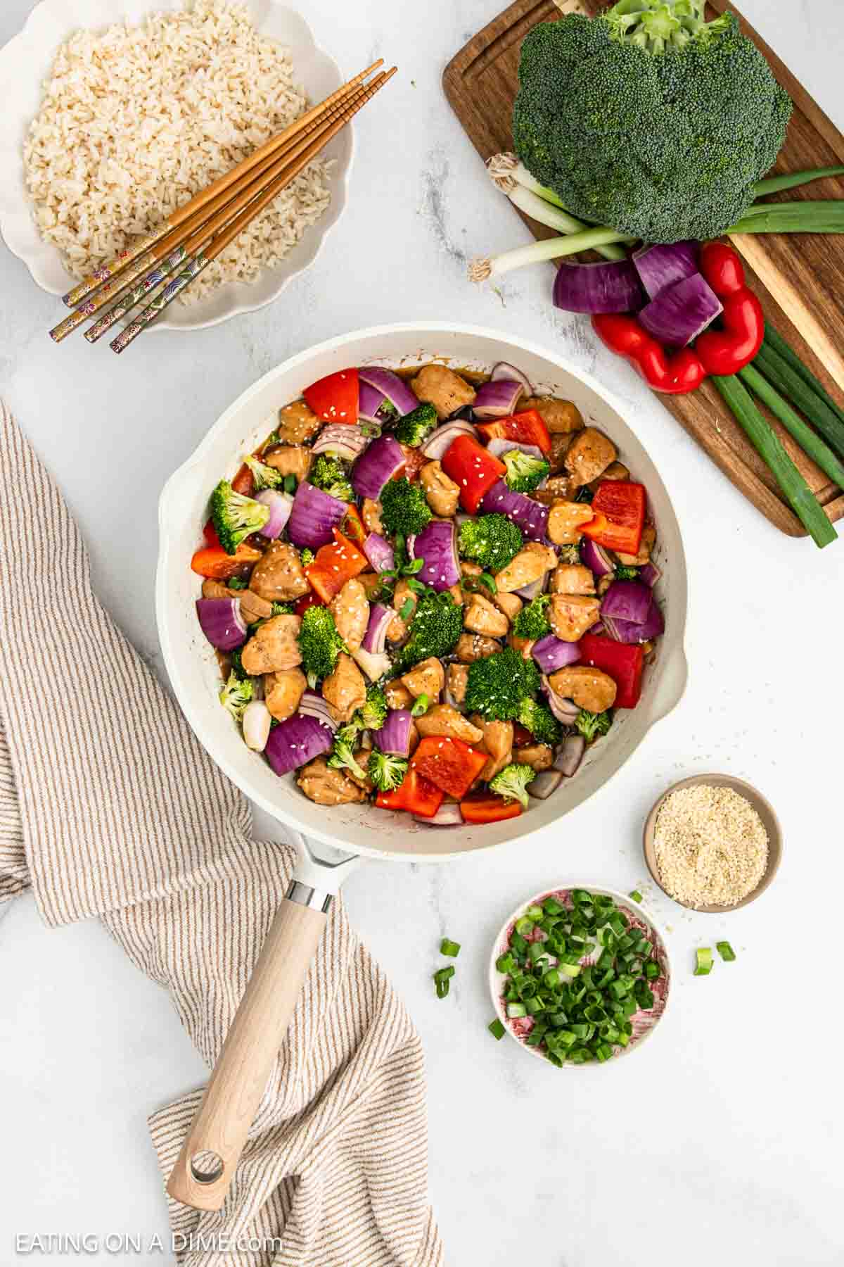 Cooked chicken, red onion, red bell pepper, and broccoli in a skillet with small bowls of chopped green onion, sesame seeds, and rice on the side.