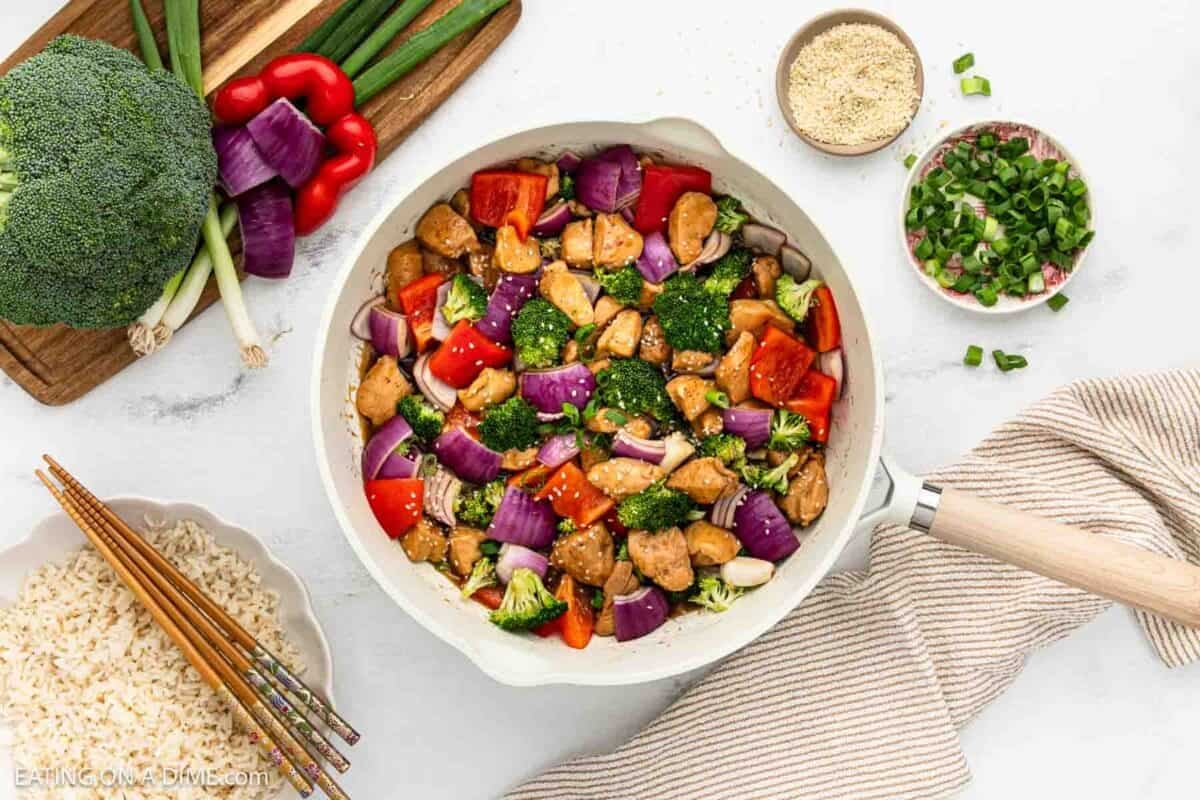 Teriyaki Chicken with broccoli, red peppers, and red onions topped with sesame seeds in a skillet. Bowl of brown rice, sesame seeds and chopped green onions on the side.