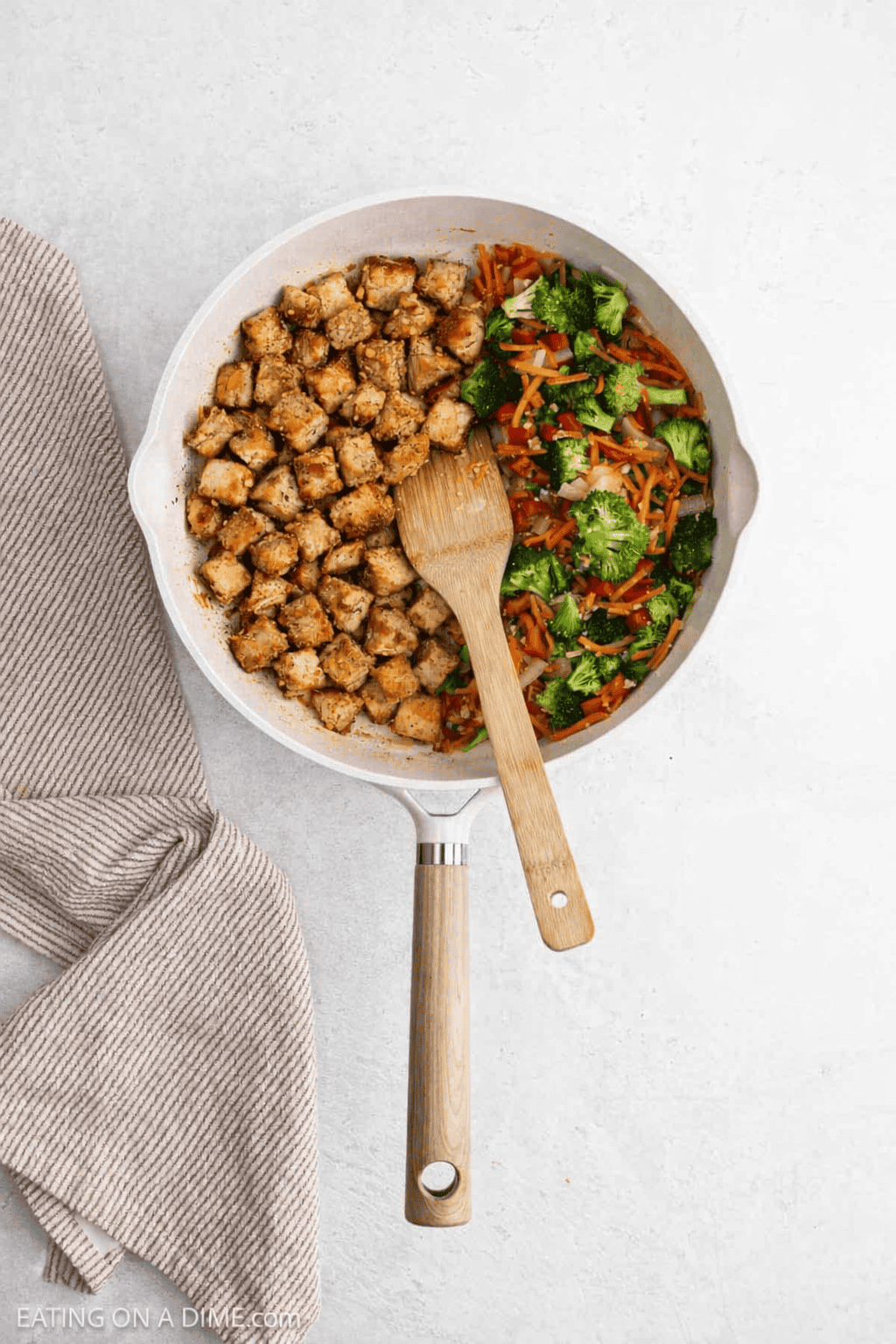 A skillet with cubed cooked chicken on one side and a mix of broccoli and shredded carrots on the other, ready for Chicken Ramen. A wooden spatula rests in the middle, with a striped cloth placed next to the skillet.