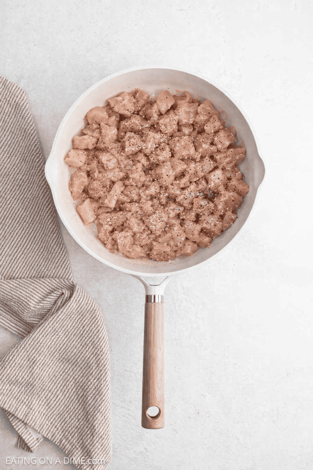 A white skillet filled with raw, diced chicken seasoned with pepper sits on a light surface next to a beige striped kitchen towel—perfectly prepped for tasty Chicken Ramen.